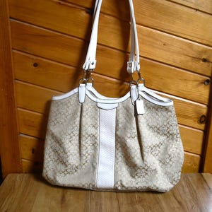 May include: A beige and tan shoulder bag featuring a repeating monogram pattern, white leather trim, two white shoulder straps, a vertical white textured centre panel, and a small white leather hang tag attached to a silver-toned metal ring.