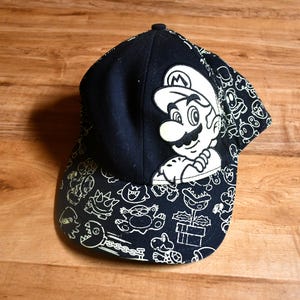 Puede incluir: Gorra de béisbol negra con un gran personaje de Mario blanco y crema en la parte delantera. La visera y los laterales están decorados con contornos blancos de personajes de dibujos animados. La gorra es un diseño clásico de seis paneles.