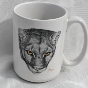 May include: White ceramic mug featuring a detailed illustration of a cougar's face. The cougar's fur is depicted in shades of gray, with striking yellow eyes. The mug has a curved handle and a glossy finish.