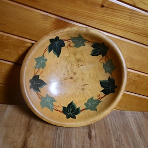 May include: A round, wooden bowl with a light brown finish. The bowl is decorated with a green and dark green ivy leaf design around the rim. The bowl is sitting on a wooden surface.