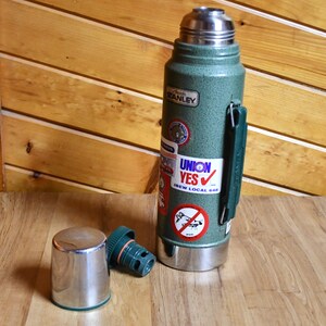 May include: A green Stanley thermos with a silver lid and base, and a green handle. The thermos has several stickers, including one that says "UNION YES". The lid and a separate green stopper are next to the thermos.