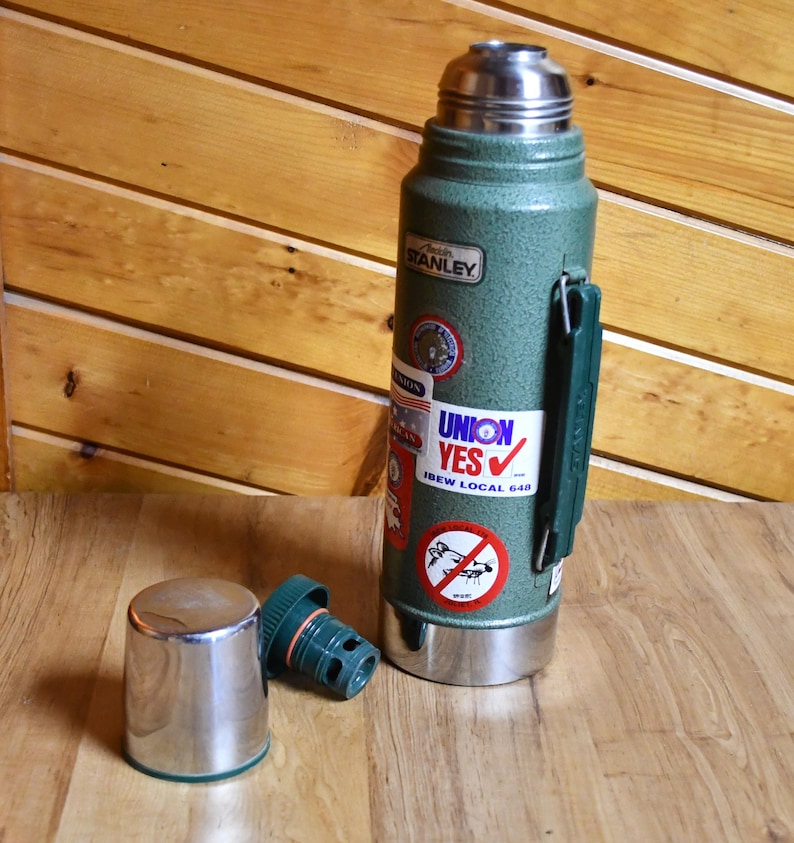 May include: A green Stanley thermos with a silver lid and base, and a green handle. The thermos has several stickers, including one that says "UNION YES". The lid and a separate green stopper are next to the thermos.