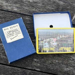 Peut inclure: Une boîte bleue de cartes à jouer "Big Deals" de taille jumbo avec une étiquette "Souvenir". La boîte est ouverte, révélant un jeu de cartes avec un bord jaune et un paysage urbain de Las Vegas. Les cartes sont sur une surface en bois vieilli.