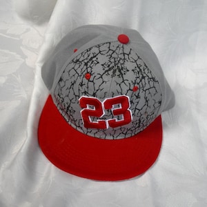 Op de afbeelding: Grijze baseballcap met een rode klep en een rode knoop. De cap heeft het nummer "23" in rood, met een gebarsten patroon op de kroon.