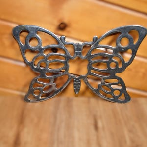 May include: A silver-colored butterfly-shaped trivet with an openwork design. The trivet is made of metal and has a detailed pattern of cut-out shapes. The wings are symmetrical, and the body has a small, pointed tail. The background is a wooden surface.