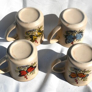 May include: Four beige ceramic mugs with curved handles, each decorated with a different butterfly design in shades of yellow, blue, red, and orange. The mugs are arranged on a white surface.