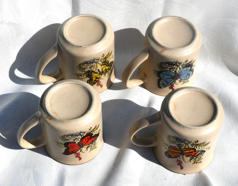 May include: Four beige ceramic mugs with curved handles, each decorated with a different butterfly design in shades of yellow, blue, red, and orange. The mugs are arranged on a white surface.