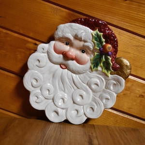 May include: A ceramic Santa Claus face plate with a white beard and rosy cheeks. The Santa's hat is burgundy with white trim. A sprig of holly with berries adorns the side of the hat. The plate is a festive holiday decoration.