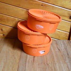 May include: Three orange plastic containers stacked on top of each other. Each container has a lid with a ridged design and a decorative floral pattern. The containers are on a wooden surface.