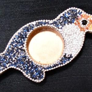 May include: A toucan-shaped trinket box made of beads. The toucan is blue, white, and yellow with a black eye. The box has a round opening in the center.