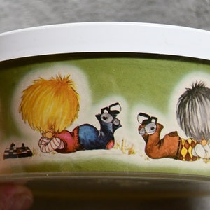 May include: A round, clear plastic container with a white lid. The container has a green background with a cartoon illustration of three children playing. The children are wearing colorful clothing and have large, expressive eyes.