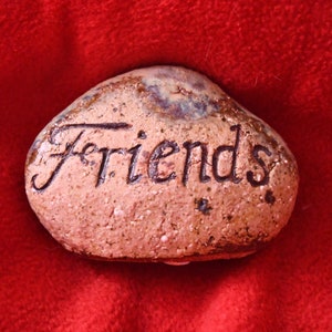 Könnte beinhalten: Ein kleiner, unregelmäßig geformter Stein mit dem Wort "Friends" in schwarzer Schrift. Der Stein hat eine gesprenkelte, hellbraune Oberfläche mit blauen und braunen Farbtupfern, vor einem leuchtend roten Hintergrund.