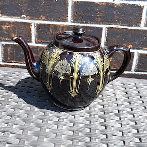 May include: A dark brown ceramic teapot featuring a glossy finish and a decorative pattern of gold-colored vertical columns and intricate filigree designs. The teapot includes a matching lid with a small knob handle and a curved spout on the side.