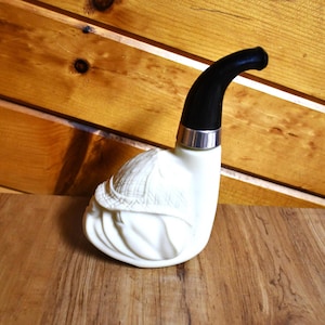 May include: A white tobacco pipe with a black curved stem and a silver band. The pipe has a detailed carved design on the bowl, resembling a hat and fabric folds. The pipe is displayed on a wooden surface.