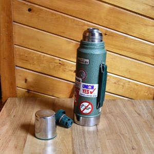 May include: A green Stanley thermos with a silver lid and handle, and a separate silver cup. The thermos has several stickers, including one that says "UNION YES". The thermos is on a wooden surface.