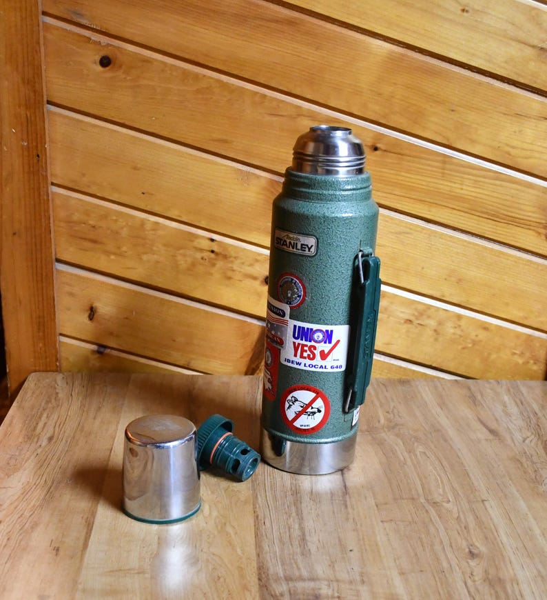May include: A green Stanley thermos with a silver lid and handle, and a separate silver cup. The thermos has several stickers, including one that says "UNION YES". The thermos is on a wooden surface.