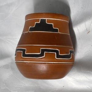 Puede incluir: Un pequeño jarrón de cerámica marrón con base redondeada y parte superior ligeramente ensanchada. El jarrón presenta un diseño geométrico en blanco y negro, con bandas horizontales y patrones escalonados. La estética general recuerda a la cerámica del suroeste.