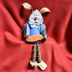 May include: A wooden bunny rabbit figurine with blue clothing, a carrot, and brown legs. The bunny has a painted face with a pink nose and brown eyes.