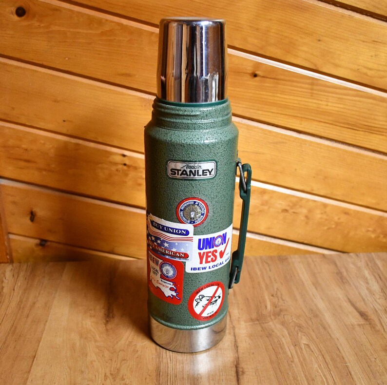 May include: A vintage green Stanley thermos with a silver lid and base. The thermos has a green handle and several stickers, including "Buy Union" and "Union Yes". The Stanley logo is visible.