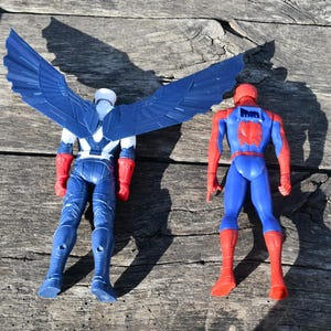 Puede incluir: Dos figuras de acci&oacute;n de superh&eacute;roes se muestran sobre una superficie de madera. Una es el Capit&aacute;n Am&eacute;rica, con un traje azul y blanco y grandes alas azules. La otra es Spider-Man, con un traje azul y rojo.