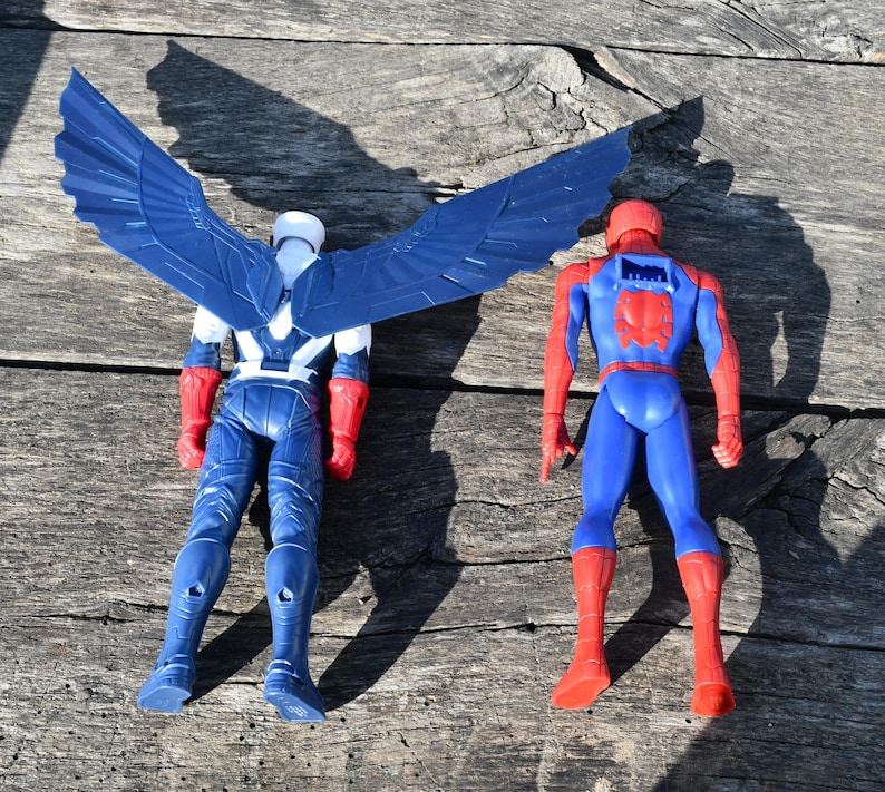 Puede incluir: Dos figuras de acci&oacute;n de superh&eacute;roes se muestran sobre una superficie de madera. Una es el Capit&aacute;n Am&eacute;rica, con un traje azul y blanco y grandes alas azules. La otra es Spider-Man, con un traje azul y rojo.
