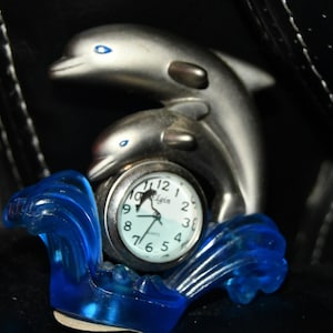 Op de afbeelding: Een zilveren klok met een blauwe golfbasis en twee dolfijnfiguren. De wijzerplaat van de klok is wit met zwarte cijfers en wijzers. De klok zegt "Elgin Quartz".