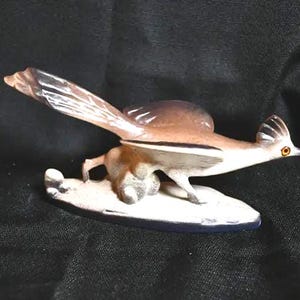 May include: A ceramic roadrunner figurine in a dynamic running pose with wings extended. The bird features brown, white, and black plumage, a prominent crest on its head, and a bright orange eye. It is mounted on a white, oval-shaped base.