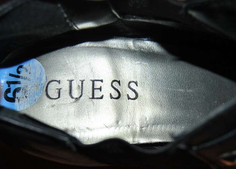 May include: Close-up of the inside of a black shoe with a silver insole that says "GUESS" in black lettering.