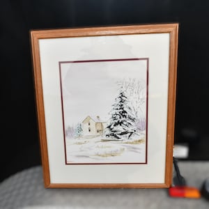 May include: Watercolor painting of a snow-covered landscape with a house and evergreen tree. The artwork is framed in a light brown wooden frame with a white mat and a maroon border. The painting depicts a winter scene.