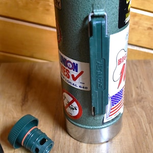 May include: A green Stanley thermos with a green handle and a silver base. The thermos has several stickers, including one that says "UNION YES" and another with an American flag design. The lid is off and lying on the wooden surface.