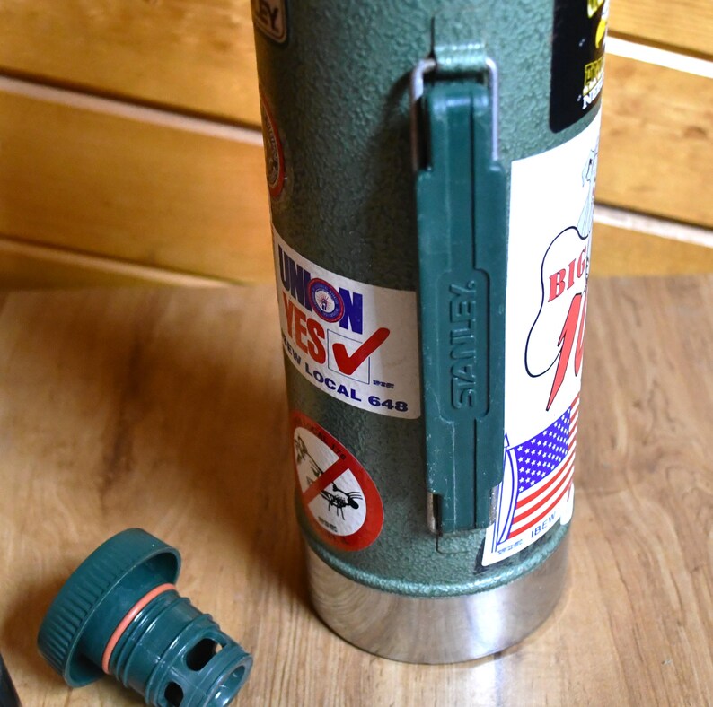 May include: A green Stanley thermos with a green handle and a silver base. The thermos has several stickers, including one that says "UNION YES" and another with an American flag design. The lid is off and lying on the wooden surface.