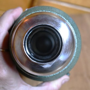 May include: Close-up view of a stainless steel and green thermos bottle. The bottle's opening is stainless steel, with a wide, circular rim. The body of the thermos is green. The image is taken from a top-down perspective.