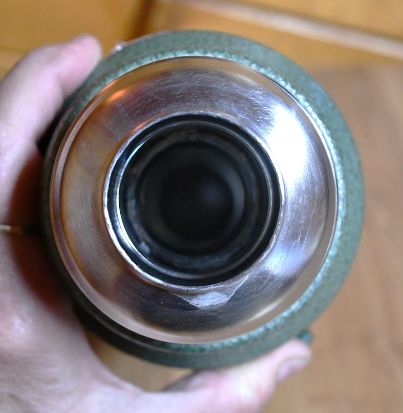 May include: Close-up view of a stainless steel and green thermos bottle. The bottle's opening is stainless steel, with a wide, circular rim. The body of the thermos is green. The image is taken from a top-down perspective.