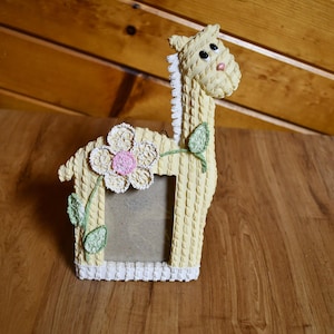 May include: A giraffe-shaped picture frame with a yellow textured finish. The frame features a large flower with a pink center and green leaves. The giraffe has black eyes and a white mane. The frame is approximately 7 inches tall.