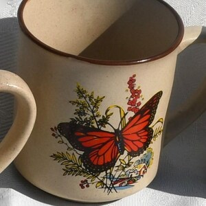 May include: Beige ceramic mug with a brown rim and handle. The mug features a vibrant illustration of a monarch butterfly with red and black wings, surrounded by greenery and flowers. A small house is also depicted.