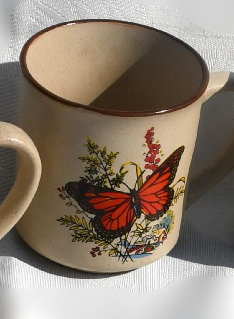 May include: Beige ceramic mug with a brown rim and handle. The mug features a vibrant illustration of a monarch butterfly with red and black wings, surrounded by greenery and flowers. A small house is also depicted.