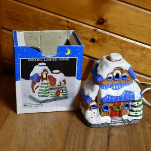 以下が含まれることがあります： 雪の屋根と青いアクセントが付いたセラミック製のライトアップハウス。箱の隣にあります。家には赤いドア、緑の花輪、小さなクリスマスツリーがあります。箱には「CERAMIC LIGHTED HOUSE」と書かれています。