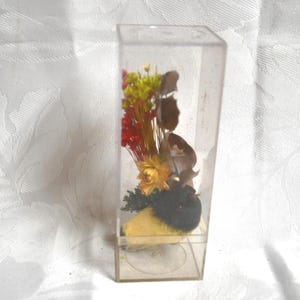 May include: A clear, rectangular plastic display case containing a dried floral arrangement. The arrangement includes yellow, red, and brown dried flowers, with a yellow and black bird-shaped element at the base. The case sits on a white, patterned surface.