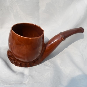 Könnte beinhalten: Eine braune Keramikpfeife-förmige Tasse mit glänzendem Finish. Die Tasse hat eine abgerundete Schale und einen gebogenen Stiel mit dekorativem Sockel. Das Design ähnelt einer traditionellen Rauchpfeife.