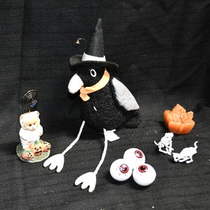 May include: A collection of Halloween decorations on a black background. Includes a black crow wearing a witch hat and orange scarf, a teddy bear figurine, a pumpkin-shaped candle, three eyeball decorations, and two small white animal figurines.