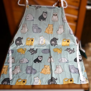 May include: A light blue apron with a white trim and a pattern of cartoon cats in various poses. The cats are in shades of gray, yellow, and white.