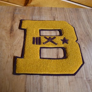 May include: A yellow felt letter "B" with a purple border, featuring a textured surface. The letter has two cut-out sections and is decorated with purple symbols including three lines, crossed bats, and a star. The patch is on a wooden surface.