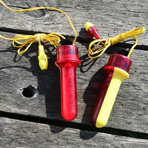 Puede incluir: Dos silbatos de plástico rojo y amarillo con cordones amarillos. Un silbato es rojo liso, el otro es mitad rojo y mitad amarillo. Los silbatos están sobre una superficie de madera envejecida.