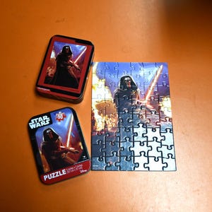 Puede incluir: Un rompecabezas de Star Wars de 50 piezas con Kylo Ren y un sable de luz rojo. El rompecabezas está en una caja de metal con una imagen de Kylo Ren en la parte delantera.