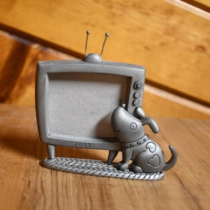 May include: A silver-colored picture frame shaped like a vintage television set, with a small dog figurine sitting beside it. The frame has a textured base and the year "2003" is inscribed on the frame.