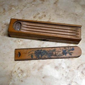 Puede incluir: Un estuche de lápices de madera vintage con tapa corredera y una regla decorada separada. La caja es rectangular con una parte superior acanalada. La regla es larga y estrecha, con un diseño floral.