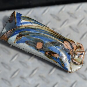 May include: A cylindrical ceramic object with a glossy finish, featuring a marbled pattern of blue, white, and brown tones. The piece has an open end and a slightly irregular, textured surface with visible glaze variations and earthy brown spots.