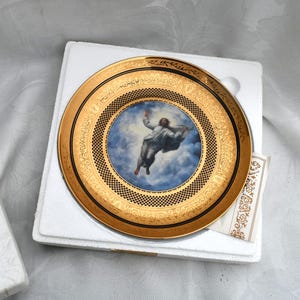 May include: A decorative plate featuring a gold rim and a central image of a figure ascending into a cloudy sky. The plate has a gold border with intricate designs and is presented in a white box.