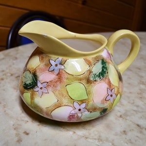 Puede incluir: Una jarra de cerámica amarilla con un diseño floral con flores rosas, verdes y moradas. La jarra tiene un cuerpo ancho y redondeado y una asa.