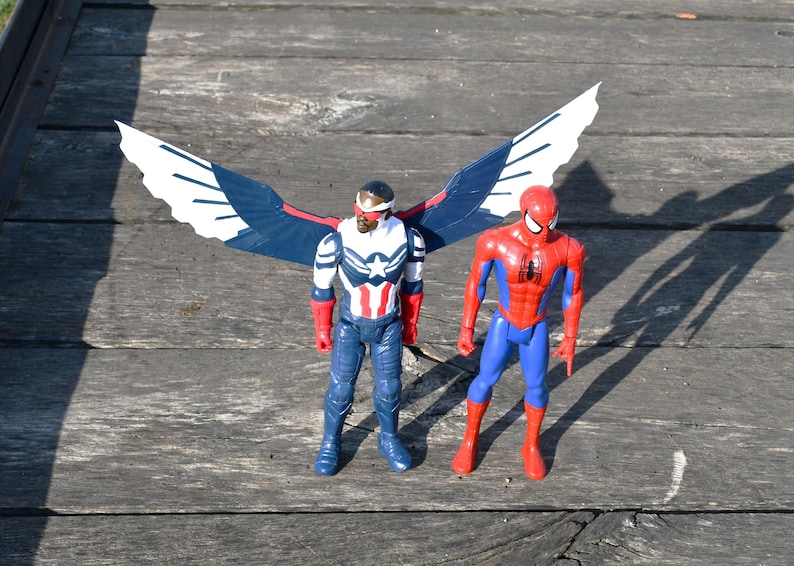 Puede incluir: Dos figuras de acci&oacute;n de superh&eacute;roes. Una figura viste un traje rojo, blanco y azul con grandes alas. La otra figura viste un traje rojo y azul. Ambas figuras est&aacute;n sobre una superficie de madera desgastada.